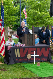 A medium shot of an outdoor Memorial Day ceremony. A man in a dark suit jacket, light blue button-down shirt, and glasses stands at a wooden podium speaking into a microphone. The podium sits on a table draped with a black tablecloth featuring the large red letters "VFW" and the text "VETERANS OF FOREIGN WARS." An American flag stands to the left. Behind the speaker, a veteran in a dark suit and a military sidecap stands with his head bowed in prayer. In front of the table, on a small patch of artificial green turf, sits a small white cross and two bronze military grave markers. Green trees and a bus stop sign are visible in the background.