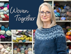 A portrait of Carol Briggs Dragos, owner of Dyed In The Wool, wearing a blue patterned knitted sweater and round glasses. She is standing in front of white cubby shelves filled with colorful skeins of yarn in various textures and shades.