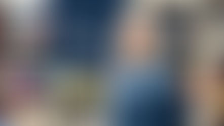 A portrait of Carol Briggs Dragos, owner of Dyed In The Wool, wearing a blue patterned knitted sweater and round glasses. She is standing in front of white cubby shelves filled with colorful skeins of yarn in various textures and shades.