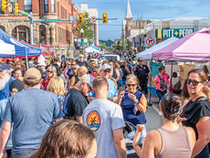 A wide-angle, sunlit photograph capturing a densely crowded street at the Greensburg Night Market. The scene is filled with people browsing between vendor tents, including a blue tent on the left and a pink tent on the right. In the foreground, a diverse crowd walks along the street. Key figures include a man in a blue "Southern Tide" t-shirt, a woman in a navy blue dress holding a beverage, and a woman in a black top and sunglasses looking at the camera. The street is lined with brick buildings, one of which has a sign reading "W Pittsburgh St." A white church steeple is visible in the distance against a blue sky. Street lights and traffic signals are present.