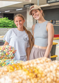 Two young women are smiling at the camera from behind a market stall counter. The woman on the left wears a grey t-shirt with "Laurel Mountain Kettle Corn" text and has long brown hair. The woman on the right wears a grey ribbed tank top, beige shorts, and a beige "Ariat" baseball cap, with a small cross necklace. In the immediate foreground, out of focus, are large bags of kettle corn. The stall is set up in front of a brick building with an "EXIT" sign and industrial garage doors. A green bush is visible to the left.
