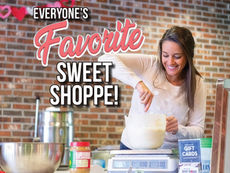 A medium shot photograph inside a sweet shop with a brick wall background, featuring a smiling woman with brown hair stirring batter in a white mixing bowl on a metal counter. Text overlay at the top reads "EVERYONE'S" followed by a large, script "Favorite" and block letters "SWEET SHOPPE!". Various kitchen items, including a large metal bowl, a peanut butter jar, a scale, and gift cards, are on the counter. Some paper heart decorations hang on the brick wall.