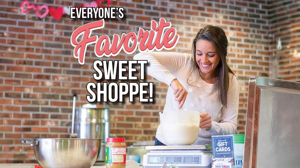 A medium shot photograph inside a sweet shop with a brick wall background, featuring a smiling woman with brown hair stirring batter in a white mixing bowl on a metal counter. Text overlay at the top reads "EVERYONE'S" followed by a large, script "Favorite" and block letters "SWEET SHOPPE!". Various kitchen items, including a large metal bowl, a peanut butter jar, a scale, and gift cards, are on the counter. Some paper heart decorations hang on the brick wall.