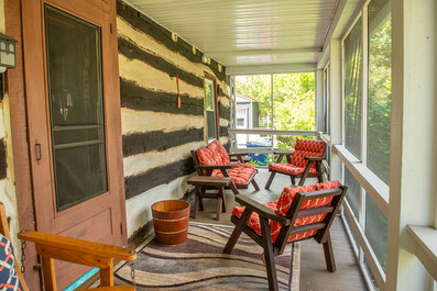 A screened-in porch with wooden furniture, featuring chairs with orange cushions, a small side table, and a log cabin-style wall. A striped rug sits on the wooden floor and a door with a screen leads into the house.