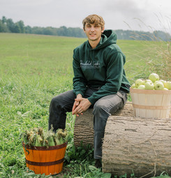 A young man wearing a dark green hoodie with the "Greenfield Basket Manufacturing, LLC" logo sits on a large, weathered log in a grassy field. To his right, a light-wood basket filled with apples sits on the log. To his left, another wooden basket, this one with orange-painted bands, is filled with harvested corn on the cob, resting on the grass.