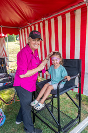 A woman in a pink shirt and baseball cap is doing face painting for a smiling young girl in a chair under a red and white striped tent.