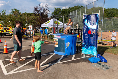 A boy throws a ball at a target on a dunk tank at a festival. A person is sitting inside the cage of the dunk tank.