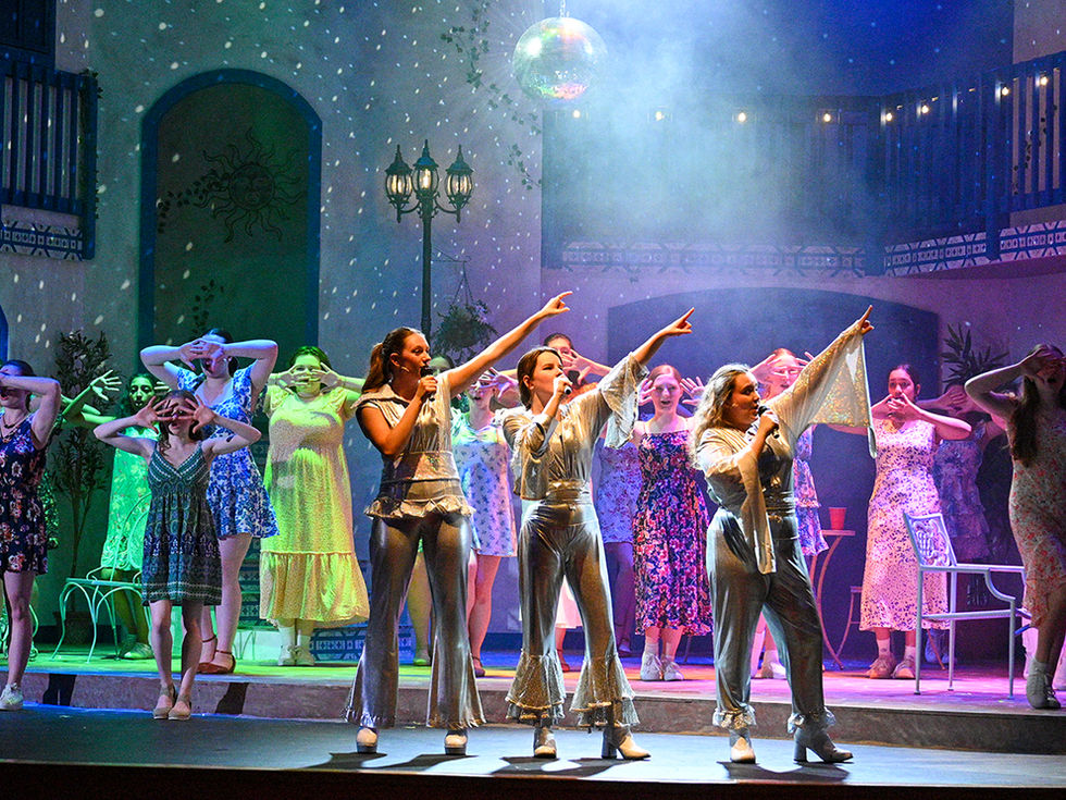 A wide stage shot featuring a large cast of young women performing in a musical, set against a Mediterranean-style village backdrop. Three central performers, dressed in matching metallic silver disco-style jumpsuits with flared legs, stand at the front, each holding a microphone and pointing one arm triumphantly toward the sky. They are illuminated by a disco ball overhead, creating a starry, shimmering effect on the stage. Behind them, other cast members in colorful floral dresses are arranged across the stage, many with their hands raised near their faces, contributing to the energetic, choreographed performance.