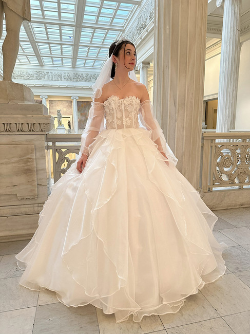 A young woman posing in a grand, marble-pillared hall wearing an intricate white wedding dress. The gown features a strapless bodice adorned with 3D floral lace, sheer off-the-shoulder sleeves, and a voluminous, multi-layered ruffled skirt. She is wearing a tiara and veil, looking off to the side.