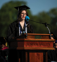 A close-up shot of a young female graduate with long brown hair and glasses giving a speech. She is wearing a black cap and gown with blue and white honor cords. She stands behind a polished wooden podium engraved with the words "SENECA VALLEY," speaking into a microphone with a bright blue foam windscreen under clear daylight.