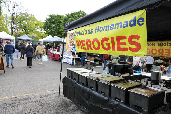 An outdoor market scene features a prominent food stall on the right under a black canopy tent, displaying a large yellow banner that reads "Delicious Homemade PIEROGIES" in red and blue letters, complete with a small photo of pierogies. Below the banner, a long table covered in black sheeting holds a series of metal chafing dishes with food. To the right, a person in a black apron works behind the counter near a large pot. The background is a bustling market aisle with people walking, other vendor stalls (including one with a visible "HUMMUS" sign), and mature trees. The ground is asphalt.
