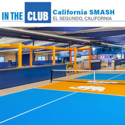 A wide-angle interior photograph of an indoor pickleball club. A large blue and orange logo graphic is overlaid in the top center. The club features multiple pickleball courts with blue playing surfaces and orange non-volley zones, separated by black nets. Court one features a prominent white bear silhouette logo. To the left, there is a lounge area with light-colored couches and low tables. Yellow overhead structures frame the lounge. The facility has a high ceiling with blue acoustic panels and exposed ductwork, with wood paneling and blue padding on the walls.