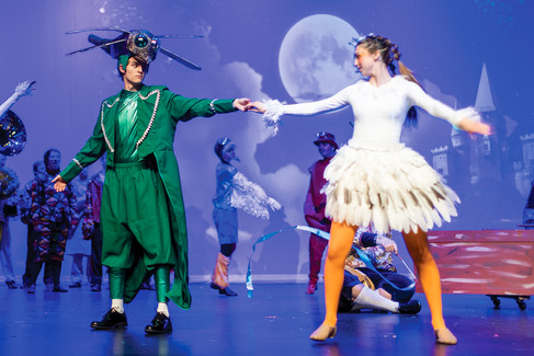 performer dressed as a dragonfly, in a green coat and insect-headpiece, dances on stage holding hands with a performer dressed as a bird, who wears a white leotard, a feathered skirt, and bright orange tights, against a background featuring a large full moon.