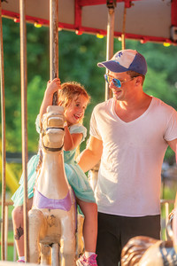 A father stands next to his young daughter who is riding a horse on a brightly lit carousel at the festival carnival.