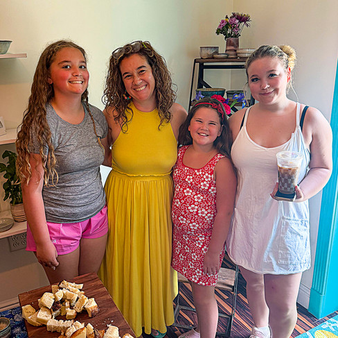 A group of four smiling people (three children and one woman) pose for a photo in what appears to be a studio or shop. They are arranged around a wooden charcuterie board piled with bread cubes. In the background are shelves with ceramic items and flowers. One child holds an iced coffee. The lighting is warm and natural.
