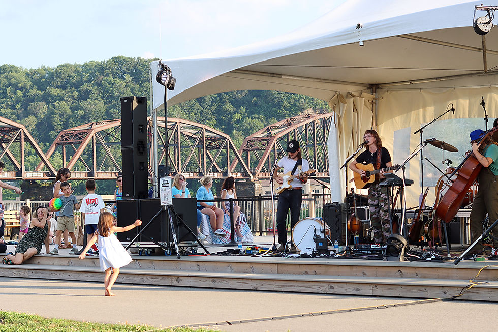 A medium, eye-level shot captures an outdoor musical performance taking place on a raised wooden stage under a large white tent. On stage, two musicians perform: one plays an electric guitar while wearing a white t-shirt and black baseball cap, and another plays an acoustic guitar while singing into a microphone, wearing a black shirt and camo-print pants. Beside them, another musician plays a double bass. In the foreground, a young girl in a white dress dances barefoot on the paved path in front of the stage. Nearby, other audience members sit and stand, including a woman in a dark patterned dress resting on the ground and two young boys, one holding a colorful ball. The background features a large, rustic-looking metal bridge spanning over a river, with lush, green, tree-covered hills rising behind it. The scene conveys a relaxed, community-focused outdoor event.