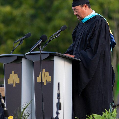 medium profile shot of a man of East Asian descent speaking at a podium during an outdoor graduation ceremony. He is wearing a black academic gown and cap, paired with a bright blue and gold master's or doctoral hood. He looks down at his notes on a dual lectern that features the gold "NA" North Allegheny logo. The background consists of soft, blurred green trees.