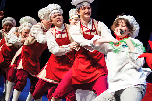 A lively scene on stage featuring a lead chef with a mustache and a group of kitchen staff in white hats and red aprons, who are playfully interacting and pulling at a green net.