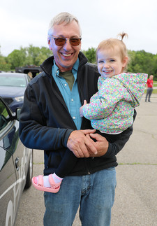 Outdoors on an asphalt lot, a senior man with grey hair and sunglasses, dressed in a black fleece jacket over a blue and olive shirt and blue jeans, smiles broadly as he holds a young girl. The girl, in a floral hooded jacket and black pants, smiles at the camera, wearing one pink sandal. The back of a blue car and trees are in the background, with an indistinct figure standing further away.