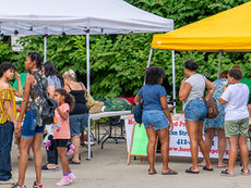 A lively crowd of shoppers and families browse outdoor vendor tents at the market on a sunny day.