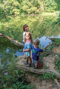 A woman and two children stand on the rocky bank of a pond. The woman is in the back, holding a fishing pole and looking at the camera. A girl with blonde hair and a boy with glasses stand in front, with fishing equipment nearby.