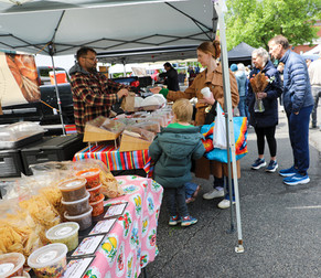 A market vendor in a red-and-black plaid flannel shirt serves a customer at his stall, which is covered with a colorful, multi-striped tablecloth. He is handing a paper bag to a woman wearing a tan trench coat, who is accompanied by a small child in a green jacket. The table is filled with various plastic containers of dips, salsas, and bags of tortilla chips. Other shoppers browse in the background under the market canopy.