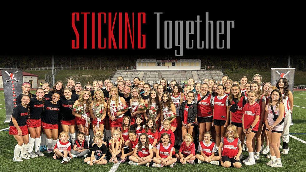 A team of female field hockey players in red and white uniforms is standing on a grassy field, posing for a group photo.