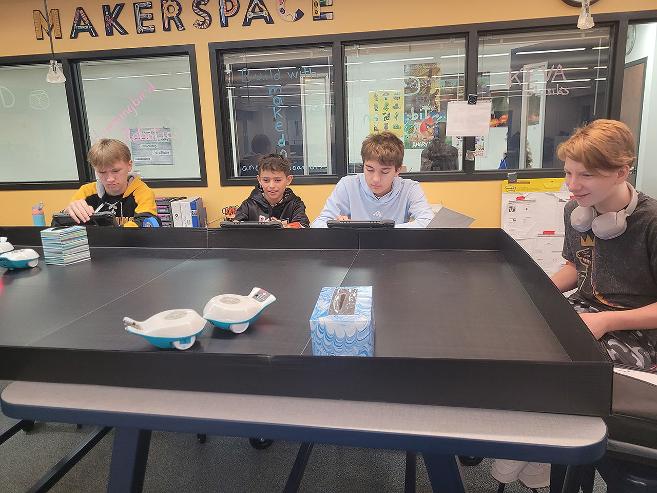 Four male middle school students collaborating around an indoor miniature robotics field in the makerspace, with small white and blue robots on the mat. They are gaining experience in Industrial & Engineering Technology through robotics.
