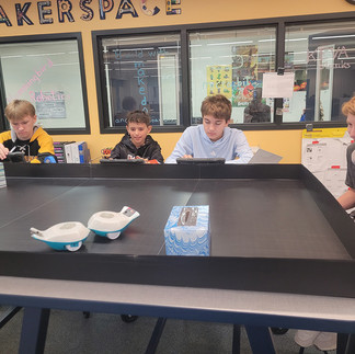 Four male middle school students collaborating around an indoor miniature robotics field in the makerspace, with small white and blue robots on the mat. They are gaining experience in Industrial & Engineering Technology through robotics.
