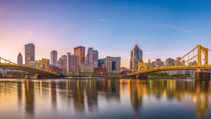 A wide panoramic view of the Pittsburgh skyline at twilight, featuring the yellow Three Sisters bridges spanning the Allegheny River. The city's high-rise buildings are reflected in the calm water as the sky transitions from soft pink to deep blue, illustrating the architectural resilience and riverside history of neighborhoods like Lawrenceville and the North Side.