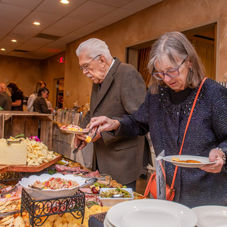 Gemini said An older woman and man serving themselves from a large buffet filled with various appetizers and a prominent cheese board at the 17th Annual Symphony of Food fundraising event.