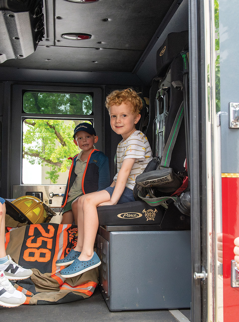 Two young boys sit inside the cab of a fire engine, exploring the firefighter seating and equipment during a community event.