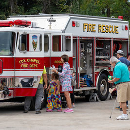 A Fox Chapel Fire Department fire rescue truck is parked with its side compartments open. A group of adults and children are gathered around the truck, looking inside the compartments and speaking with personnel.