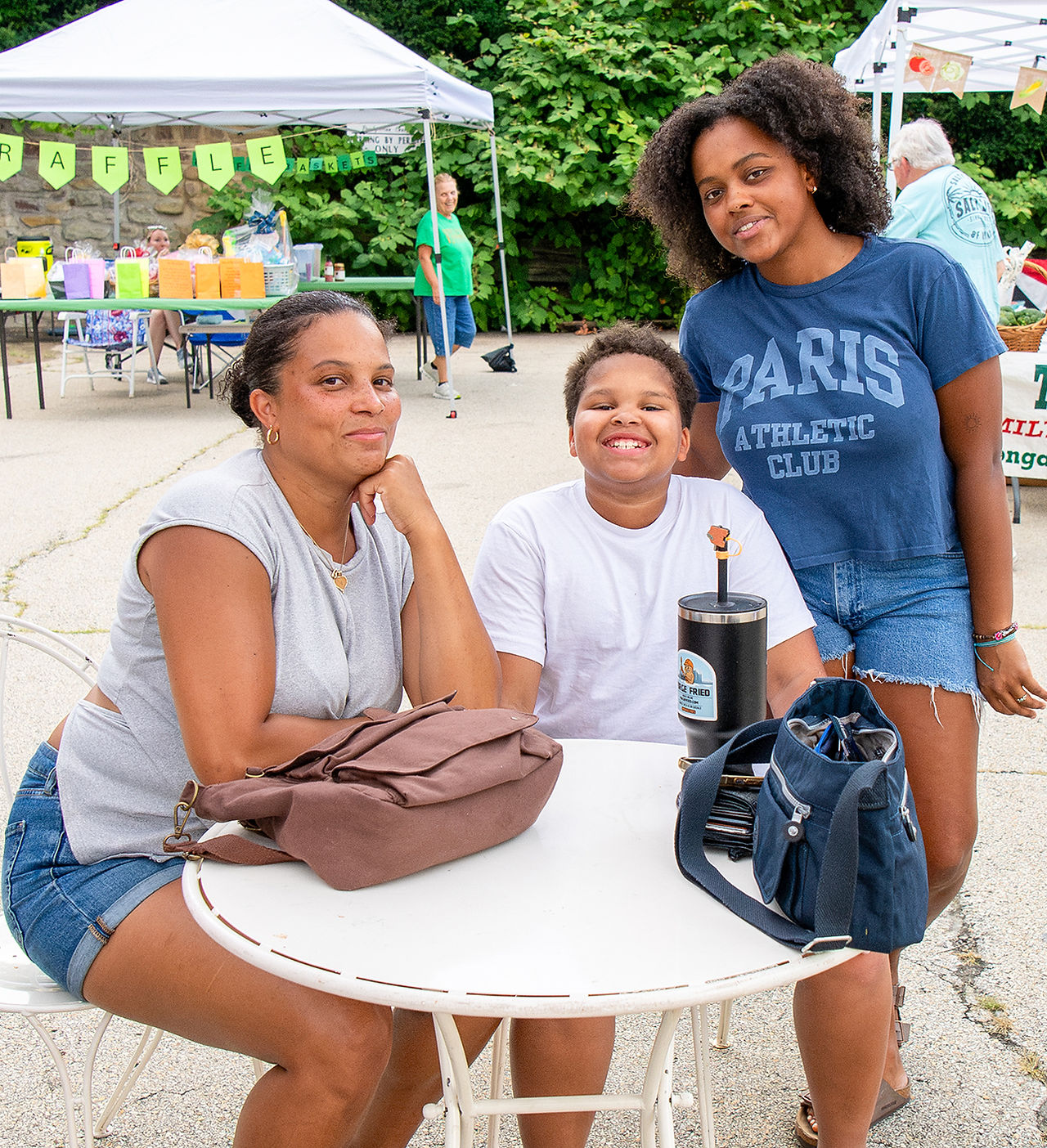 hree people sit together at an outdoor white table, smiling and posing for the camera at the market.
