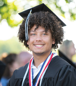 A close-up portrait of a smiling male graduate with a prominent curly afro hairstyle. He is wearing a black mortarboard cap, a black graduation gown, and red, white, and blue honor cords around his neck over a white collared shirt. The background is softly blurred with green trees and sunlight filtering through.