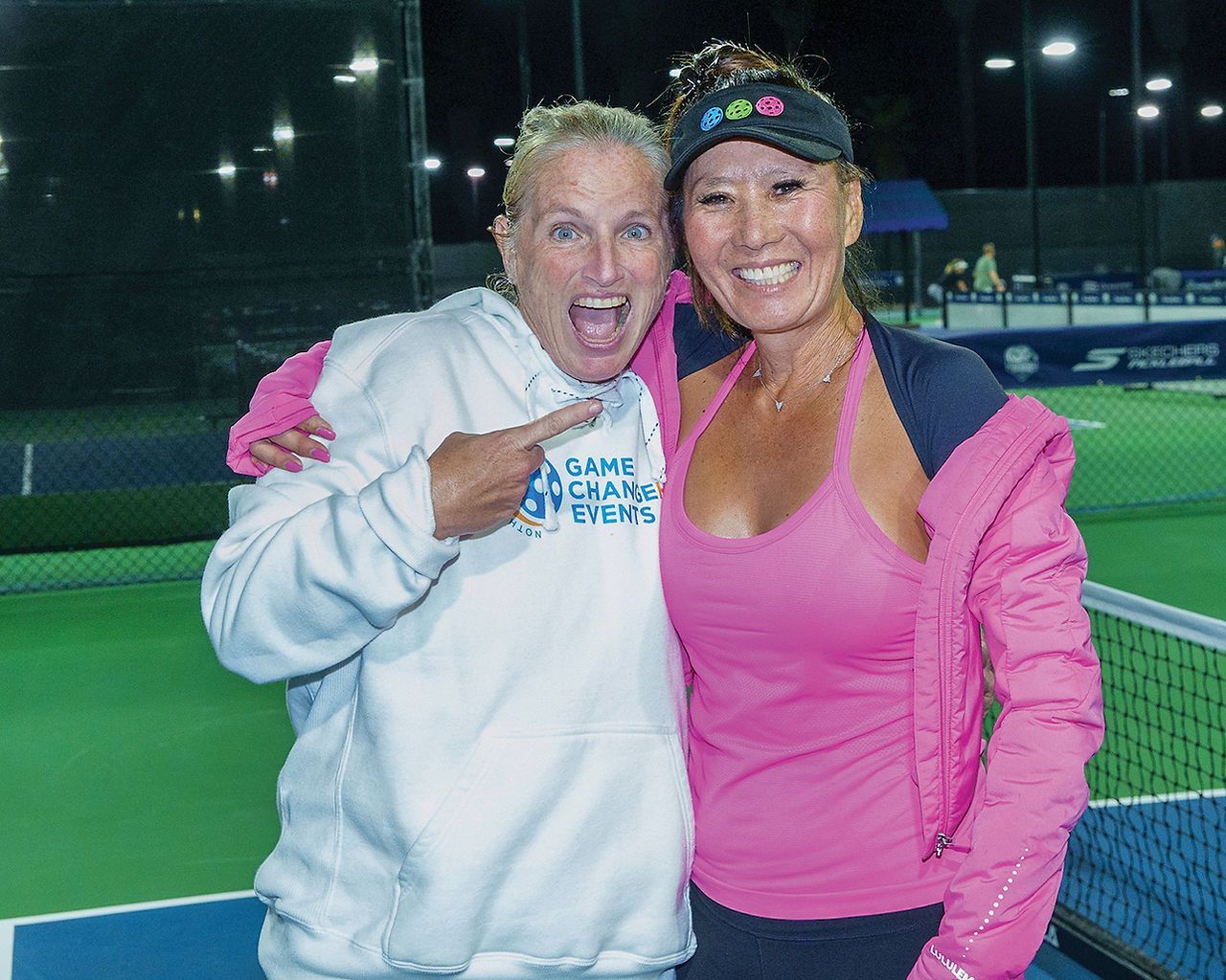 Pro Women's Doubles gold medal winners Lee Whitwell (left), wearing a white hooded sweatshirt, and Jennifer Dawson (right), wearing a pink tank top and black visor, embrace and smile on the court.