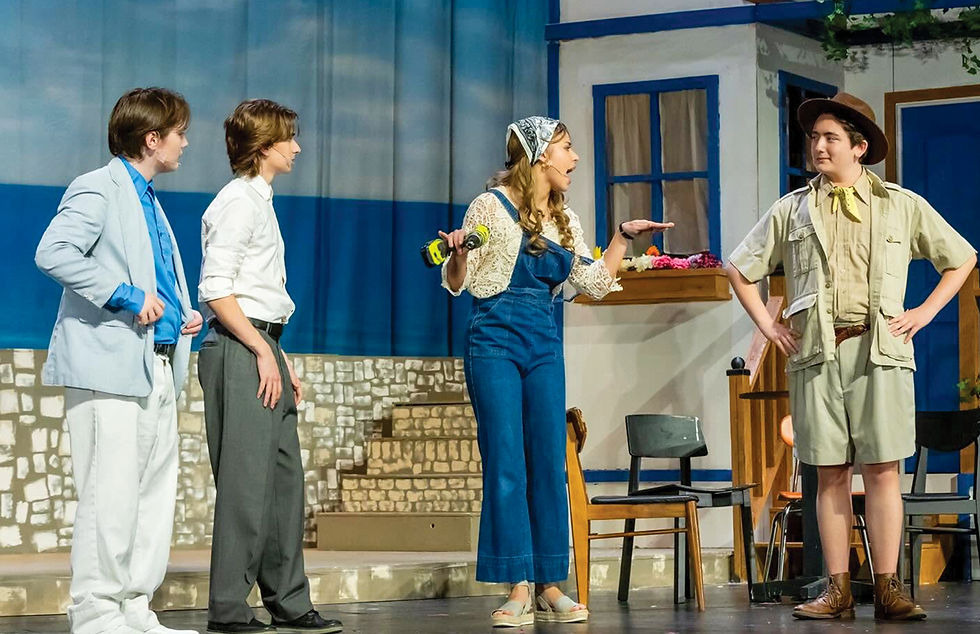 A medium shot of four actors performing a scene on a theater stage. On the far left, an actor in a light-blue jacket, white shirt, and white pants looks toward the center. Next to him, an actor in a white button-down shirt and grey trousers also looks toward the center. In the middle, an actress with a headscarf, crocheted top, and denim overalls holds a drill and gestures while speaking loudly. On the far right, an actor dressed as a safari guide in a tan shirt, shorts, and hat stands with hands on hips, listening. The stage features the same stone-block steps and beach backdrop seen in the crew photos, but also includes a house front with a blue window and door. A wooden chair and a black modern chair are placed near the center. The floor is a dark stage surface.