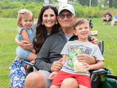 A multi-generational family—a woman, an older man, and two young children—smile warmly while sitting together in lawn chairs on a grassy field. The young boy in the foreground holds a bag of popcorn, capturing a candid moment of family fun at a community event like a Lunchtime Concert or Movie in the Moonlight at Robin Hill Park.