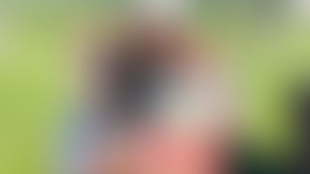 A multi-generational family—a woman, an older man, and two young children—smile warmly while sitting together in lawn chairs on a grassy field. The young boy in the foreground holds a bag of popcorn, capturing a candid moment of family fun at a community event like a Lunchtime Concert or Movie in the Moonlight at Robin Hill Park.