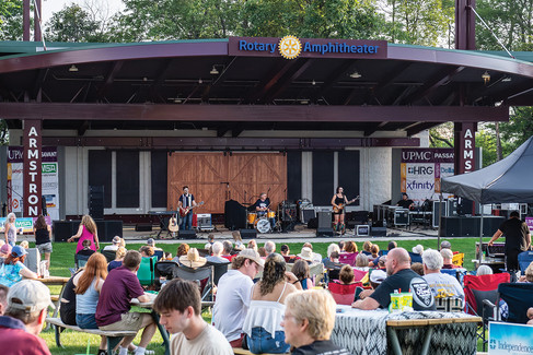 A band performs on the Rotary Amphitheater stage to an audience seated in lawn chairs and at picnic tables on a grassy field.
