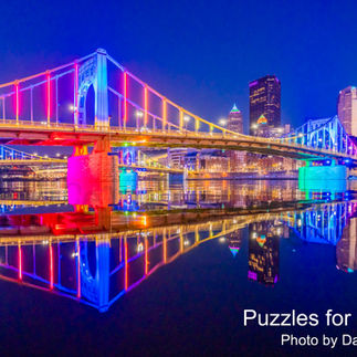 A wide photo of a bridge with large, rectangular stone piers. The span is lit by multiple colors: blue, purple, magenta, gold, and green lights. The structure is reflected on the still surface of the water, below. To the right is the dark city skyline, with one skyscraper lit by blue lights, and other buildings lit by white and yellow lights. To the left are a row of smaller buildings, reflecting in the water. At the bottom right in a white, serif font, the text reads: Puzzles for Patients. Below this in a smaller font is the text: Photo by Dave DiCello.