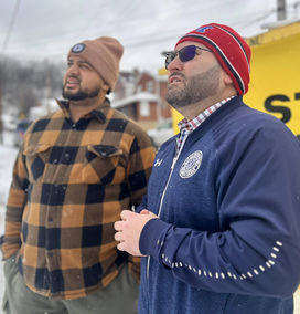 Mayor Tom Maglicco (right) stands outdoors on a snowy day alongside another man, both looking upward and to the left. The Mayor is wearing a red, white, and blue knit beanie, sunglasses, and a navy blue zip-up jacket with a circular seal on the chest over a plaid shirt. The man on the left wears a tan beanie and a brown-and-black checkered jacket. The background shows a snowy residential area with a yellow structure partially visible.