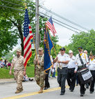 A medium shot of a six-person color guard and honor guard marching in a parade on a paved road with double yellow lines. In the front, a man on the left in a full tan desert camouflage uniform carries a large American flag. To his right, a man in a white short-sleeve shirt with a lanyard and dark trousers carries a blue flag with yellow text, parts of which read "VETERANS," "POST," and "POST 66." Behind them, another veteran in a white uniform shirt wears a dark blue cap and carries a small snare drum on a sling, which has a sign attached reading, "VIETNAM VETERANS MEMORIAL... DORMONT HIGH SCHOOL ALUM... KIA JAMES A. BARRET... JEFFREY M. WALSH... DAVID W. HENSEL." The color guard is walking under an overcast sky, with lush green trees and utility lines in the background.