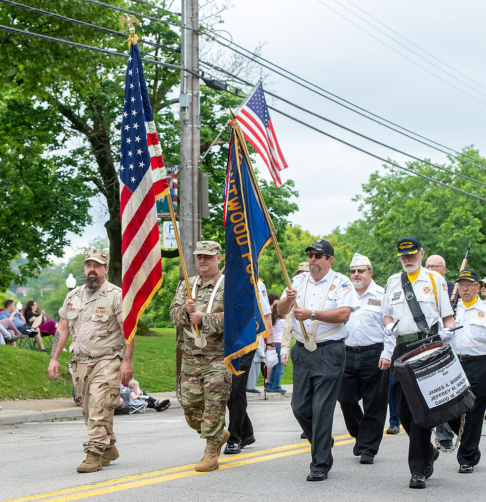 A medium shot of a six-person color guard and honor guard marching in a parade on a paved road with double yellow lines. In the front, a man on the left in a full tan desert camouflage uniform carries a large American flag. To his right, a man in a white short-sleeve shirt with a lanyard and dark trousers carries a blue flag with yellow text, parts of which read "VETERANS," "POST," and "POST 66." Behind them, another veteran in a white uniform shirt wears a dark blue cap and carries a small snare drum on a sling, which has a sign attached reading, "VIETNAM VETERANS MEMORIAL... DORMONT HIGH SCHOOL ALUM... KIA JAMES A. BARRET... JEFFREY M. WALSH... DAVID W. HENSEL." The color guard is walking under an overcast sky, with lush green trees and utility lines in the background.