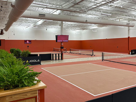 An indoor pickleball facility featuring multiple courts with red surfaces, white boundary lines, nets, and red walls. Potted ferns sit in the foreground, and digital scoreboards are mounted in the background.
