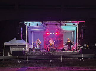 A band named Jukebox performs on an outdoor stage at night. The stage is lit with red and purple lights, and the band members are playing a guitar, keyboard, and drums.