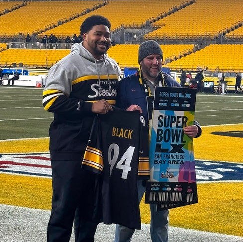 Pittsburgh Steelers defensive lineman Yahya Black (left) stands on a football field alongside pantry founder AJ Owen (right). Yahya is wearing a grey Steelers hoodie and holds his own black number 94 jersey. AJ is in a blue jacket and beanie, smiling broadly while holding two giant, colorful "SUPER BOWL LXI SAN FRANCISCO BAY AREA" commemorative tickets. Both men are looking at the tickets, with the expansive empty yellow and gold stadium seats visible in the background.