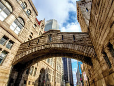 A low-angle view of the historic stone "Bridge of Sighs" archway connecting the Allegheny County Courthouse and the Old Allegheny County Jail in downtown Pittsburgh. The architectural landmark is set against a blue sky with modern city skyscrapers visible in the background.