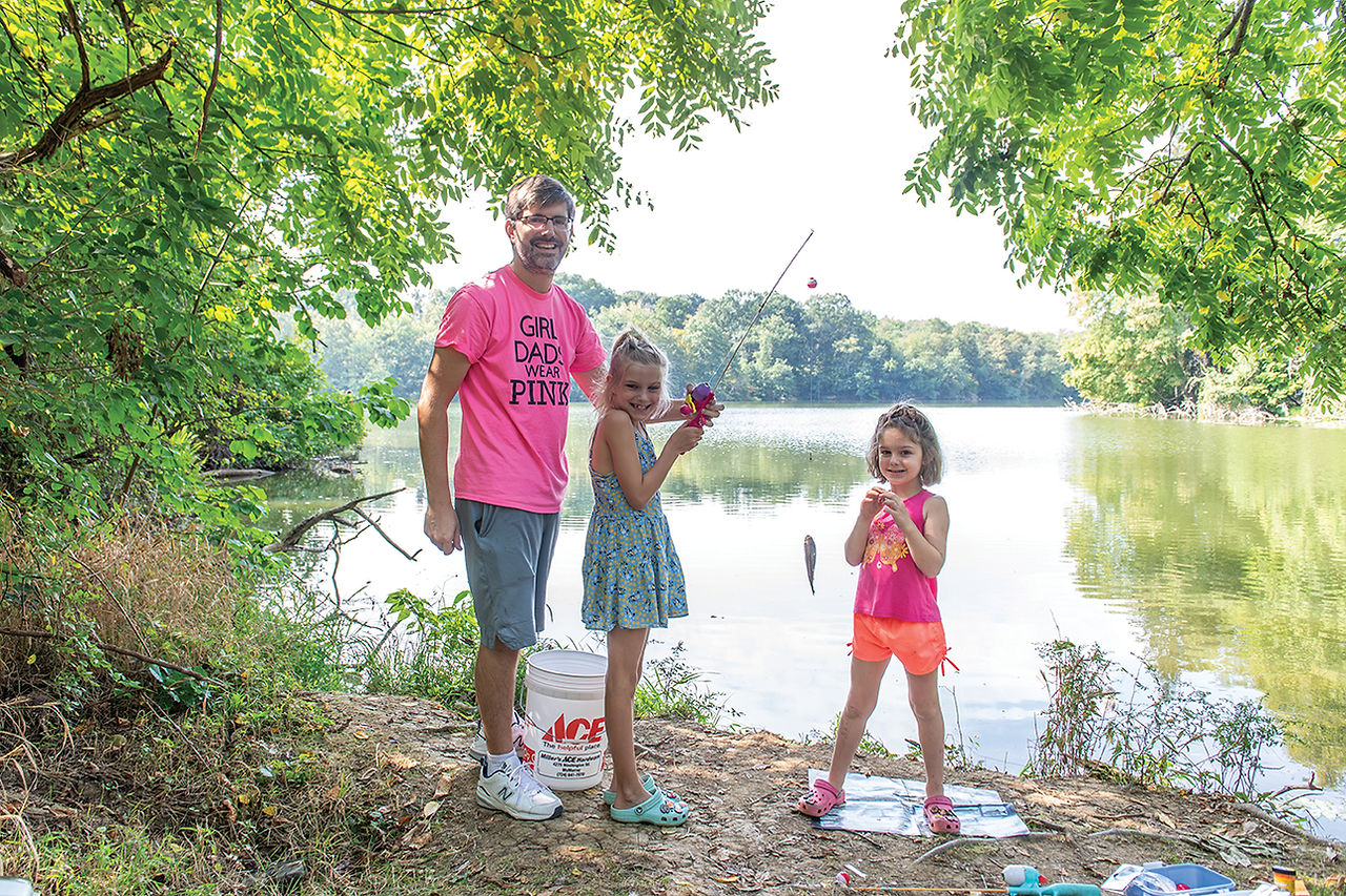 A family of three stands on the grassy bank of a pond on a sunny day. A man in a pink t-shirt smiles at the camera, a girl with a blue dress holds up her fishing rod with a fish on the line, and another girl with a pink top and shorts stands beside her, smiling.