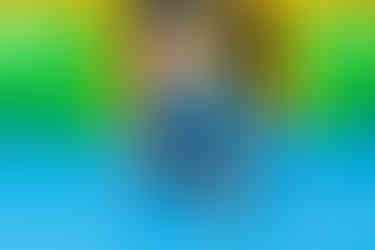 A happy toddler in a blue shirt is crawling on a bright blue inflatable bouncy surface, with the green and yellow sides of the bounce house visible.